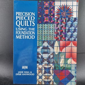 May include: A book titled "Precision Pieced Quilts Using the Foundation Method" with a blue cover. The cover features the title in white text and a grid of colorful quilt squares. The authors are Jane Hall and Dixie Haywood.