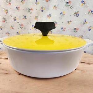 May include: White ceramic casserole dish with a yellow lid and a black knob. The dish is sitting on a wooden surface.
