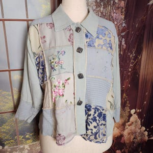 May include: A light blue patchwork jacket with floral embroidery and patterned fabric squares. The jacket features a collared neckline, long sleeves, and square, dark buttons. The patchwork includes floral and paisley designs.