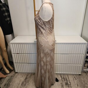 May include: A sleeveless floor-length dress in a light beige hue, featuring an intricate geometric pattern composed of metallic gold beadwork and sequins that extends from the bodice down to the hemline, creating a structured linear design.