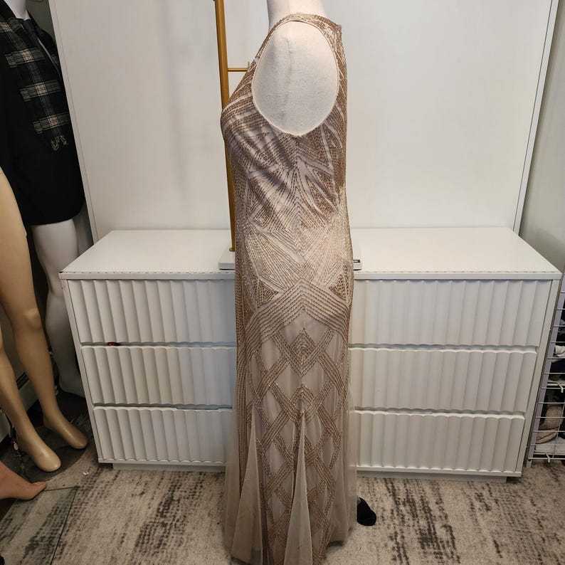 May include: A sleeveless floor-length dress in a light beige hue, featuring an intricate geometric pattern composed of metallic gold beadwork and sequins that extends from the bodice down to the hemline, creating a structured linear design.
