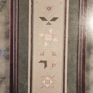 May include: A framed, vertical needlepoint piece with a repeating geometric pattern in shades of beige, brown, and pink. The frame is dark brown. The needlepoint features a series of stylized floral and geometric shapes.