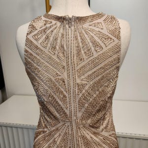 May include: A sleeveless dress featuring a sheer beige base adorned with intricate gold beadwork arranged in geometric patterns. The garment includes a central vertical zipper closure running down the back, showcasing detailed metallic embellishments.