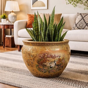 May include: A large, round, decorative ceramic planter with a detailed landscape scene, filled with tall, green snake plants. The planter has a gold and brown finish with a detailed rim and is placed on a striped rug.