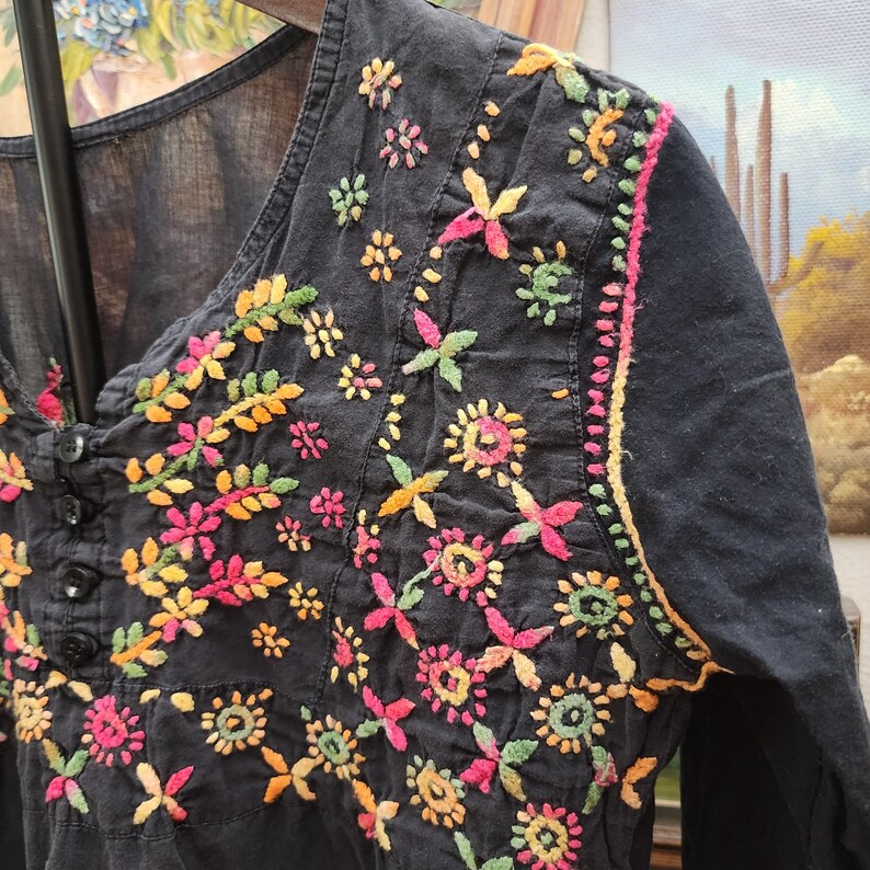 Peut inclure: Haut noir brod&eacute; avec des motifs floraux color&eacute;s. La broderie pr&eacute;sente des fleurs et des feuilles roses, jaunes, oranges et vertes. Le haut a une encolure carr&eacute;e et des boutons.