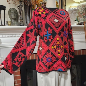May include: A red and black patterned sweater with long sleeves. The sweater features geometric designs in various colors, including yellow, blue, and white. The sweater is displayed on a mannequin.