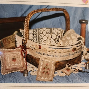 May include: A woven wicker basket with a handle, filled with lace and cross-stitch samplers. The samplers feature floral and needlework designs, with the text "Lesley Rudnicki her Sampler Basket". A small pillow and a needlework card are also visible.