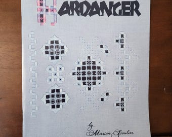 May include: A book titled "Hardanger" by Marion Scoular, featuring a cover with a black and white geometric design and a pink and white design in the upper left corner.