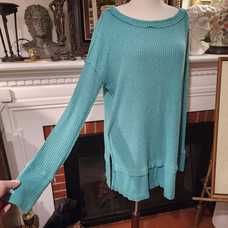 Puede incluir: Una blusa de manga larga de punto de gofre color turquesa con un cuello ancho y aberturas laterales. La blusa se muestra en un maniqu&iacute;. Las mangas tienen pu&ntilde;os acanalados. La tela tiene un aspecto texturizado.