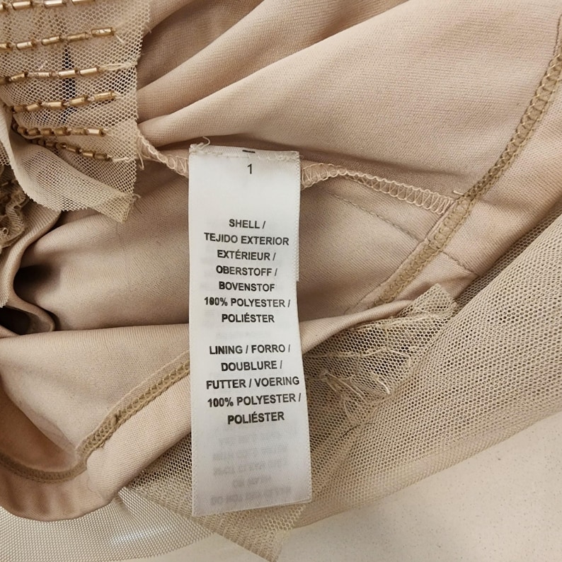 May include: A beige garment featuring a white care label with the text: SHELL / TEJIDO EXTERIOR / EXT&Eacute;RIEUR / OBERSTOFF / BOVENSTOF 100% POLYESTER / POLI&Eacute;STER LINING / FORRO / DOUBLURE / FUTTER / VOERING 100% POLYESTER / POLI&Eacute;STER. The fabric is mesh.