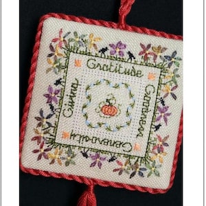 May include: Square, embroidered wall hanging with a red border and tassel. The center features a pumpkin and the words "Gratitude, Giving, Goodness, Generosity". Floral and bird designs surround the text.