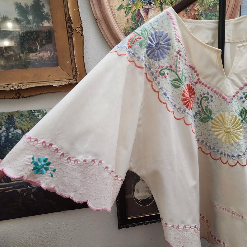 May include: Cream-colored embroidered blouse with a V-neckline and wide sleeves. The blouse features colorful floral embroidery in shades of blue, pink, yellow, and green. The sleeves are trimmed with a scalloped edge and a turquoise flower.