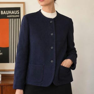 May include: A navy blue jacket with a round collar and three buttons. The jacket has two front pockets and a white turtleneck underneath. The Bauhaus poster is visible in the background.