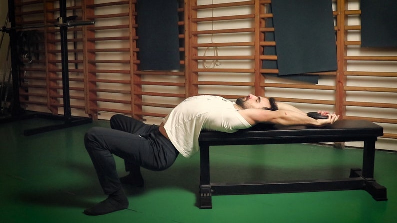 May include: A person performing a bridge exercise on a black bench, holding a weight plate. The person is wearing a white t-shirt and dark pants. The background includes wooden wall bars and a green floor.