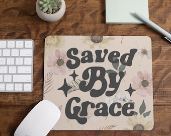 May include: A beige mousepad with the words "Saved By Grace" in dark gray, surrounded by floral and star designs. A white keyboard, mouse, pen, and sticky notes are also visible.