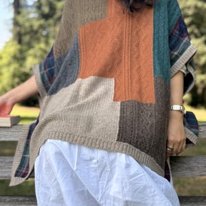 Pode incluir: Um poncho de patchwork com uma mistura de texturas de malha trançada e malha lisa em tons de bege, castanho e laranja, acentuado com painéis de tecido xadrez azul e verde. A peça tem gola alta e bainha canelada, usada sobre uma saia branca.