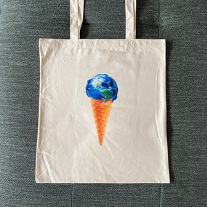 Op de afbeelding: Een witte canvas draagtas met een afbeelding van de aarde, gedrukt in de vorm van een ijshoorn. De aarde is blauw en groen, en de hoorn is oranje.
