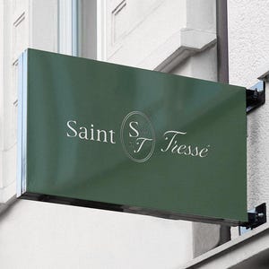 May include: A rectangular, dark green sign with the words "Saint Tresse" in white script. The sign is mounted on a white building exterior with a security camera visible. The sign is attached with black metal brackets.