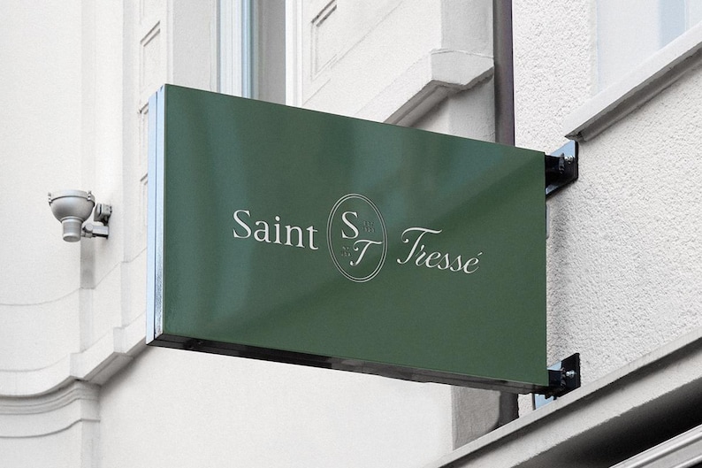 May include: A rectangular, dark green sign with the words "Saint Tresse" in white script. The sign is mounted on a white building exterior with a security camera visible. The sign is attached with black metal brackets.