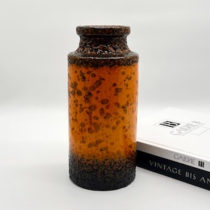 Könnte beinhalten: Eine zylindrische Keramikvase mit strukturierter, gesprenkelter Oberfläche. Die Vase geht von einem dunkelbraunen Sockel in einen leuchtend orangefarbenen Mittelteil über, mit einem strukturierten braunen Rand. Die Vase ist ein dekorativer Gegenstand.
