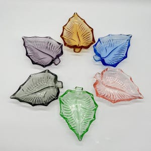 Könnte beinhalten: Eine Ansammlung von sechs blattförmigen Glasschalen, die in einem kreisförmigen Muster angeordnet sind. Jede Schale hat eine eigene durchscheinende Farbe, darunter Bernstein, Blau, Rosa, Grün, Grau und Violett, mit geprägten Aderdetails.