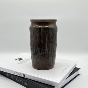 Può includere: Un vaso in ceramica marrone con linee verticali strutturate. Il vaso ha una parte superiore marrone scuro e una forma cilindrica. È appoggiato su una pila di libri con il testo "GALERIE B" e "VINTAGE BIS ANTIK".