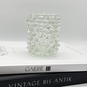 Può includere: Un portacandele in vetro trasparente con una superficie testurizzata a forma di bolle. Il portacandele è posto sopra due libri, uno con il testo "GALERIE B" e l'altro con il testo "VINTAGE BIS ANTIK".