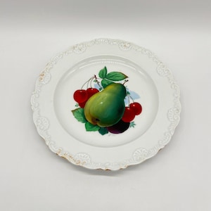 Può includere: Piatto decorativo bianco con bordo smerlato e finiture dorate. Il centro presenta una natura morta di una pera verde, ciliegie rosse e foglie verdi. Il piatto è su sfondo bianco.