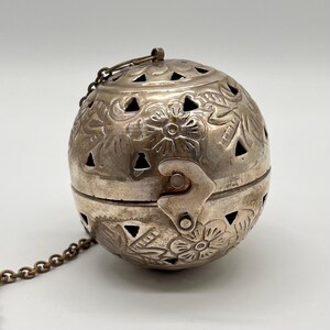 May include: A silver-colored, spherical tea infuser with floral and leaf patterns. The infuser has a hinged opening with a clasp and a small chain attached. Decorative cut-out shapes are visible throughout the design.
