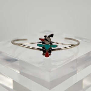 Op de afbeelding: Een zilveren manchetarmband met een kleurrijk, geometrisch ontwerp. Het centrale ontwerp is een gestileerde figuur met zwarte, rode, turquoise en witte accenten. De armband wordt gepresenteerd op een helder, schuin oppervlak.