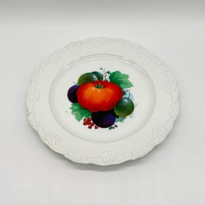 Op de afbeelding: Witte keramische plaat met een geschulpte rand. De plaat heeft een centraal ontwerp van een levendige oranje tomaat, groene en paarse vruchten en rode bessen. De rand van de plaat heeft een delicaat, reliëf bloemmotief.