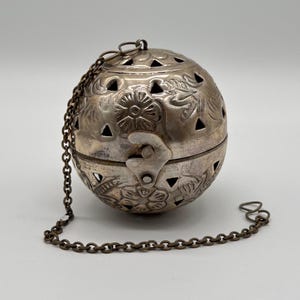Może przedstawiać: Srebrny, kulisty pojemnik z kwiatowymi i geometrycznymi wycięciami. Pojemnik posiada otwór na zawiasach i dołączony łańcuszek. Powierzchnia zdobiona jest wytłoczonymi wzorami kwiatowymi. Pojemnik służy prawdopodobnie do przechowywania drobnych przedmiotów.