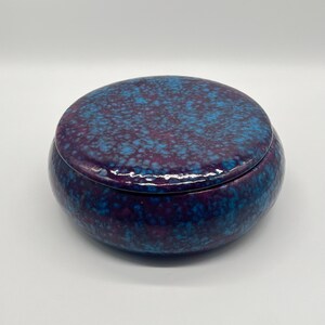 Könnte beinhalten: Eine runde Keramik-Schmuckdose mit Deckel. Die Dose hat eine tiefblaue bis violette Farbe mit einem gesprenkelten Muster aus hellerem Blau. Der Deckel passt gut auf den Boden und bildet ein zusammenhängendes und dekoratives Stück.