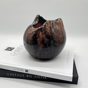 Peut inclure: Un vase en céramique marron foncé fait à la main, avec une surface texturée et une finition brillante. Le vase est posé sur deux livres avec le texte "GALERIE B" et "VINTAGE BIS ANTIK" imprimé sur les couvertures. Le vase a une forme organique unique.