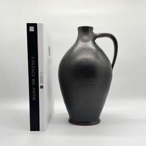 Könnte beinhalten: Ein dunkel anthrazitfarbener Keramikkrug mit bauchigem Körper, engem Hals und einem geschwungenen Henkel. Neben dem Krug steht ein weißes Buch mit schwarzem Buchrücken, auf dem der Text "VINTAGE BIS ANTIK" und das Logo "GALERIE B" stehen.