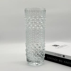 Może przedstawiać: Przezroczysty szklany wazon z teksturowanym wzorem przypominającym pęcherzyki. Cylindryczny wazon ma szeroki otwór i stoi prosto. Wazon stoi obok książki z tekstem "GALERIE B" i "VINTAGE BIS ANTI".
