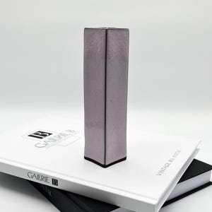 Könnte beinhalten: Eine hohe, rechteckige Keramikvase mit einer hellvioletten Glasur. Die Vase hat eine leicht strukturierte Oberfläche und einen dunkelgrauen Rand.