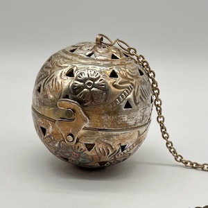 May include: A silver-colored, spherical container with intricate floral and leaf patterns. The container has triangular cutouts and a hinged clasp. A delicate chain is attached to the top. The container is likely used for decorative or functional purposes.