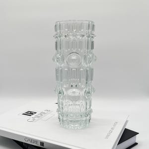 Può includere: Un vaso di vetro trasparente con un design geometrico strutturato. Il vaso ha una forma cilindrica con motivi ripetuti di cerchi e linee verticali. Il vaso è appoggiato su un libro.
