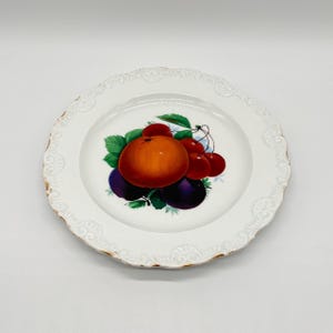 Op de afbeelding: Witte keramische plaat met een geschulpte rand en gouden rand. De plaat heeft een kleurrijke illustratie van een sinaasappel, kersen en pruimen. Het fruit is omgeven door groene bladeren.