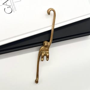 Peut inclure: Un marque-page en forme de singe de couleur laiton. Le marque-page représente un singe suspendu à un crochet incurvé, avec un corps long et fin et des traits faciaux détaillés. La queue du singe se termine par un petit pied.