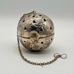 Op de afbeelding: Zilverkleurige, bolvormige thee-infuser met ingewikkelde bloemen en geometrische uitgesneden ontwerpen. De infuser is scharnierend en heeft een ketting bevestigd, eindigend in een kleine lus. Het metaal vertoont tekenen van ouderdom en slijtage.