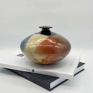 Könnte beinhalten: Eine gedrungene, abgerundete Keramikvase mit glänzender Oberfläche in den Farben Dunkelblau, Creme und gebranntes Orange. Die Vase hat einen schmalen, dunkelbraunen Hals und eine kleine kreisförmige Öffnung an der Oberseite mit glatter Textur.