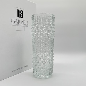 Puede incluir: Un jarrón de vidrio transparente con un patrón texturizado en forma de burbujas. El jarrón es cilíndrico y se mantiene en posición vertical. El texto "GALERIE B" y "VINTAGE BIS ANTIK" es visible en una caja blanca en el fondo.