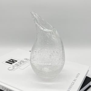 Könnte beinhalten: Eine klare, tropfenförmige Glasvase mit einem ausgestellten Ausguss. Die Vase hat eine strukturierte Oberfläche mit kleinen Luftblasen. Die Vase steht auf einem weißen Buch.