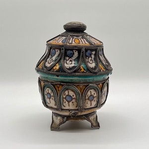 Op de afbeelding: Een keramische en zilveren pot met deksel, versierd met ingewikkelde geometrische patronen in blauw, geel en wit. De pot heeft drie zilveren voetjes en een zilveren band rond de bovenkant.