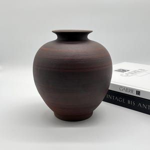 Könnte beinhalten: Eine dunkelbraune, kugelförmige Keramikvase mit matter, texturierter Oberfläche und horizontalen Streifen. Die Vase hat einen schmalen, ausgestellten Rand und einen abgerundeten Körper, der sich zu einem kleinen Boden verjüngt, neben zwei Büchern.