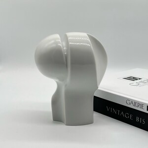 Könnte beinhalten: Eine moderne, abstrakte Skulptur aus glänzend weißer Keramik. Das Stück hat eine abgerundete Oberseite mit einem vertikalen Schlitz und eine geschwungene, organische Form. Die Skulptur steht neben einem Buch mit dem Text "GALERIE VINTAGE BIS".