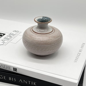 Könnte beinhalten: Eine kleine, runde Keramikvase mit geripptem, hellbeigem Körper und einem glatten, ausgestellten Hals. Das Innere der Öffnung ist mit einer dunkelblau-grau gesprenkelten Glasur versehen, während der äußere Rand und der Boden eine natürliche, unglasierte braune Tontextur aufweisen.