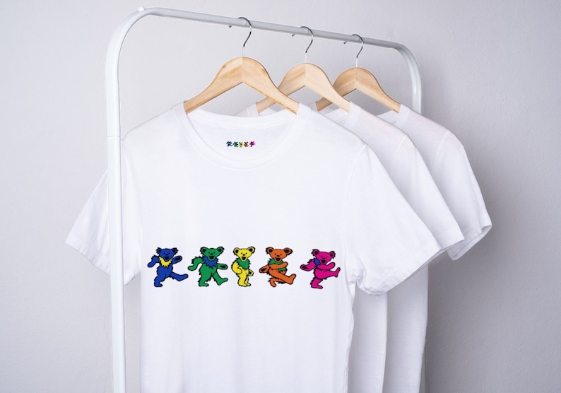 May include: Three white t-shirts with short sleeves hang on wooden hangers. The front of the t-shirt on the left features a row of five colorful dancing bears.